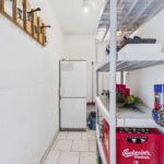 Ein schmaler Raum mit einem Kühlschrank, Regalen voller Gegenstände und einer Wand mit Garderobenhaken.