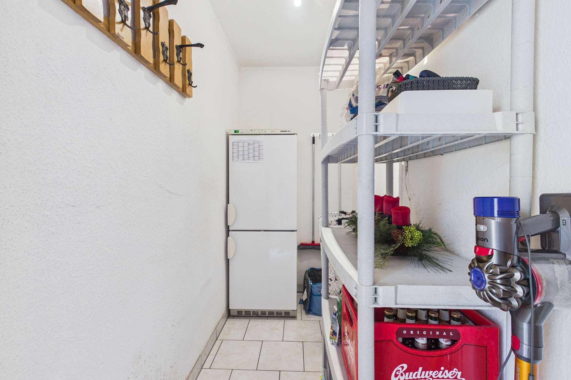 Ein schmaler Raum mit einem Kühlschrank, Regalen voller Gegenstände und einer Wand mit Garderobenhaken.