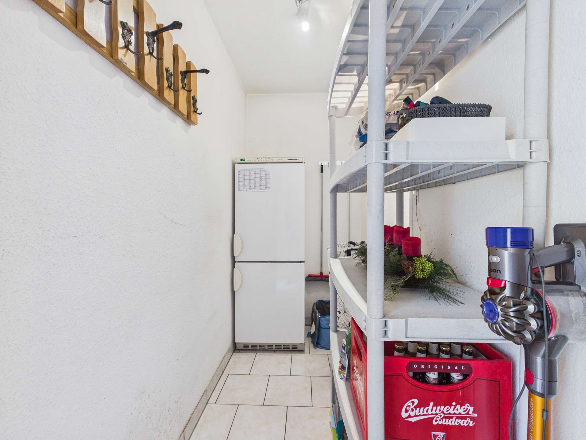 Ein schmaler Raum mit einem Kühlschrank, Regalen voller Gegenstände und einer Wand mit Garderobenhaken.