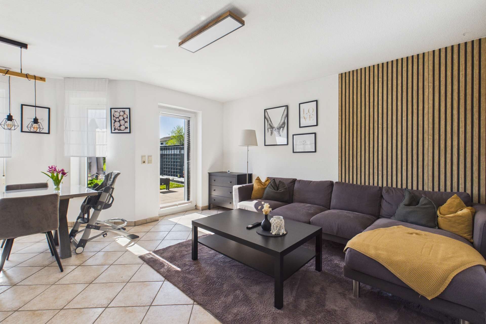 Modernes Wohnzimmer mit einer grauen Couch, einem schwarzen Tisch und einem Essbereich, hell erleuchtet durch Fenster.