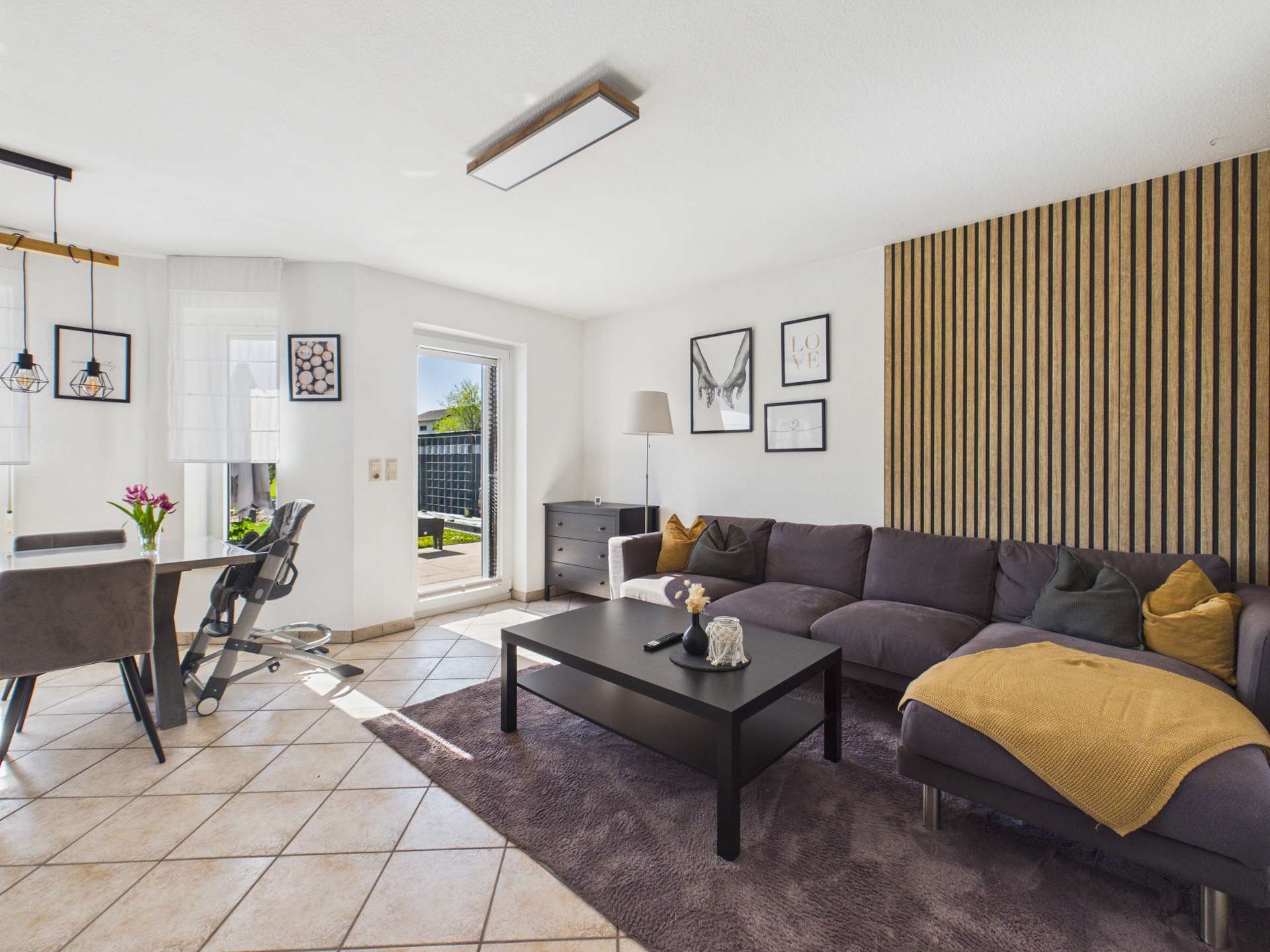 Modernes Wohnzimmer mit einer grauen Couch, einem schwarzen Tisch und einem Essbereich, hell erleuchtet durch Fenster.
