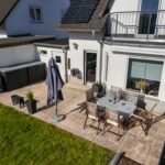 Terrasse mit Tisch, Stühlen und Sonnenschirm, umgeben von Rasen und modernen Wohngebäuden.