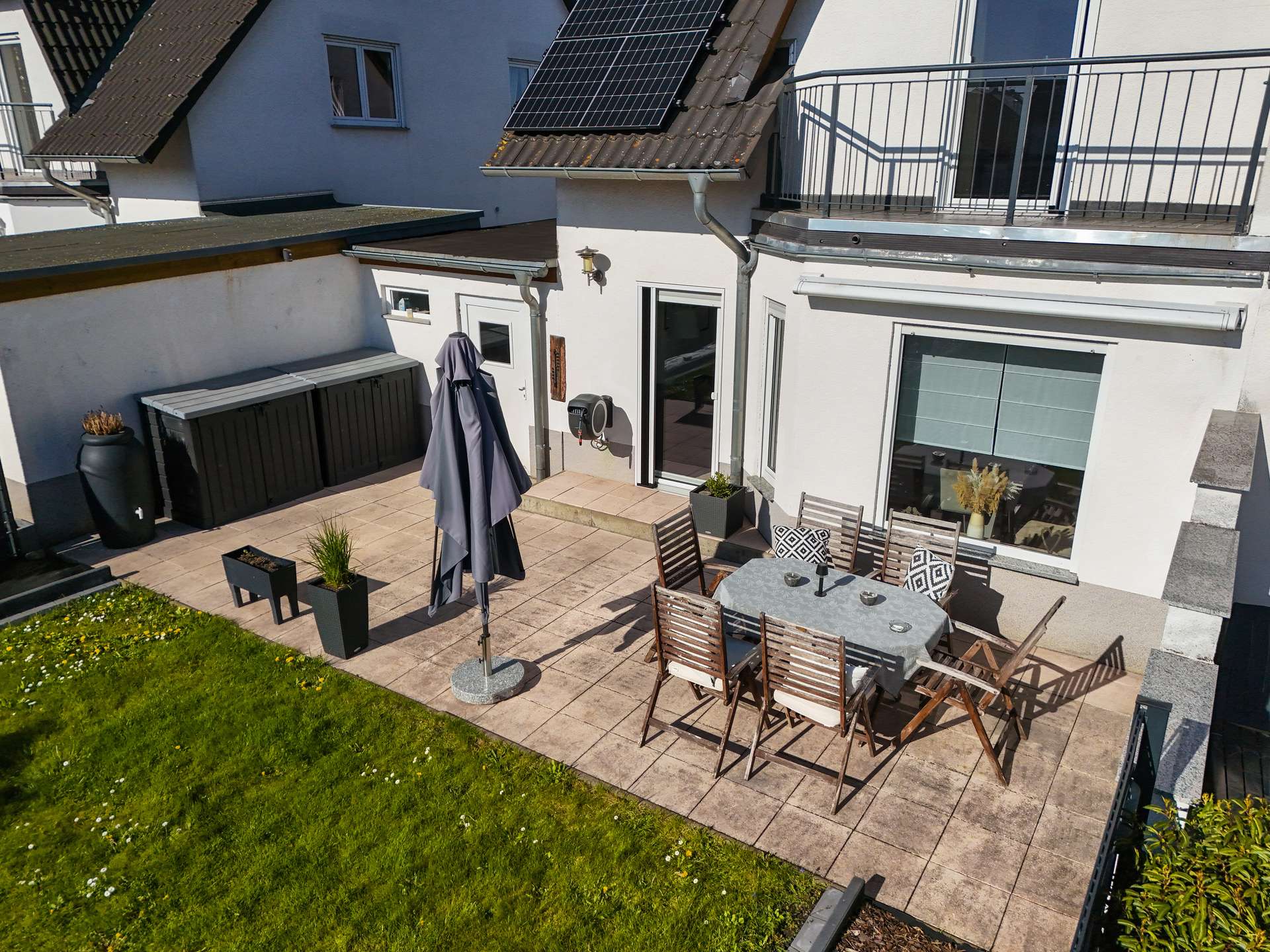 Terrasse mit Tisch, Stühlen und Sonnenschirm, umgeben von Rasen und modernen Wohngebäuden.