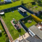 Luftaufnahme eines umzäunten Gartens mit Rasen, Pavillon, Spielplatz und verschiedenen Pflanzen.