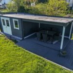 Eine moderne Gartenlaube mit einem überdachten Bereich und einem Esstisch aus schwarzen Stühlen auf einer Holzterrasse.