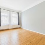 Helles, leeres Zimmer mit Holzfußboden, zwei Fenstern und grauer Wandfarbe. Vorhänge hängen an den Fenstern.