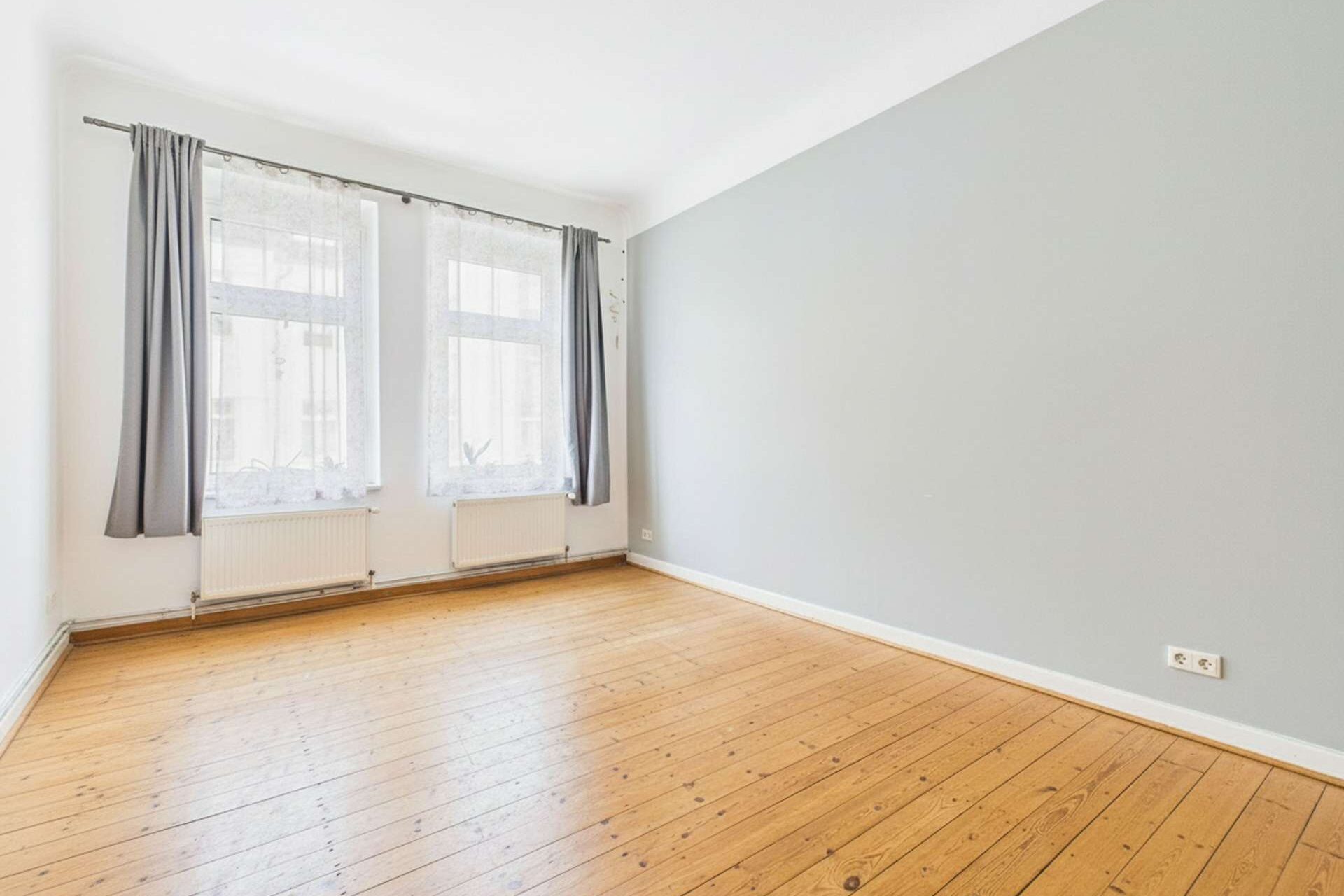 Helles, leeres Zimmer mit Holzfußboden, zwei Fenstern und grauer Wandfarbe. Vorhänge hängen an den Fenstern.