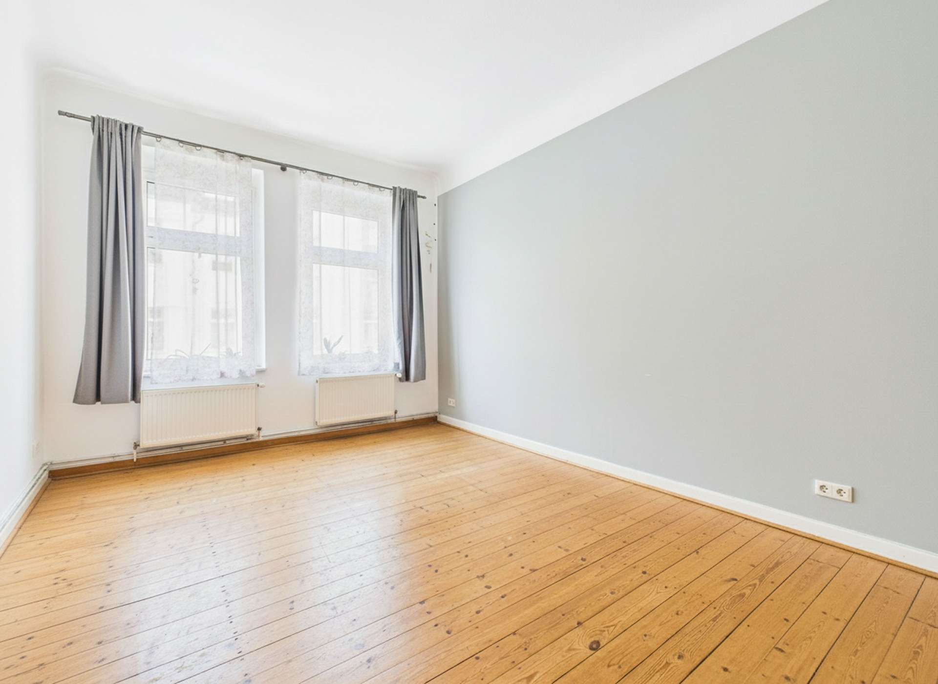 Helle, leere Wohnung mit Holzfußboden, großen Fenstern und grauer Wand. Vorhänge an den Fenstern.