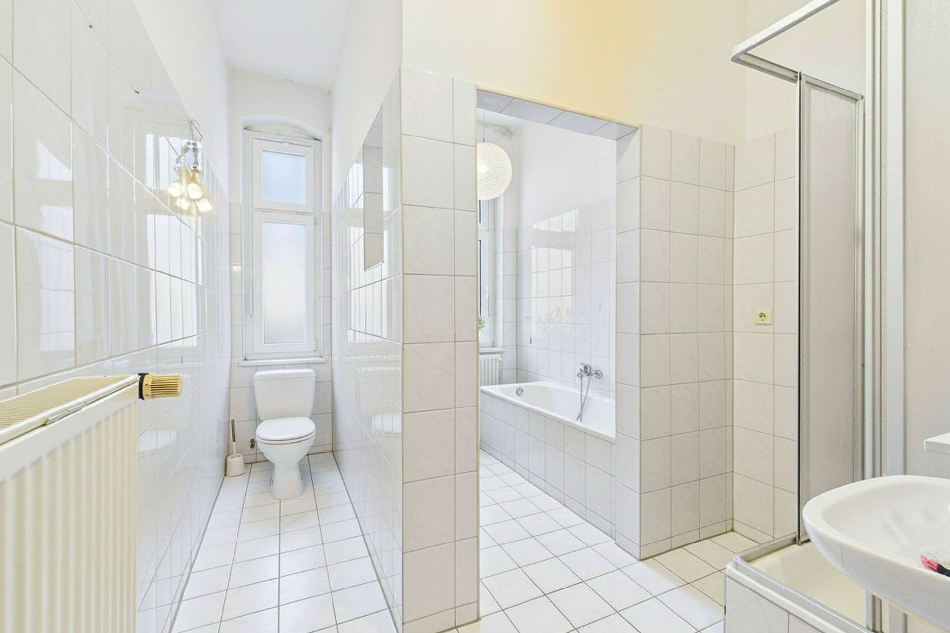 Helles Badezimmer mit weißen Fliesen, Badewanne, Dusche, WC und Fenster für natürliche Beleuchtung.