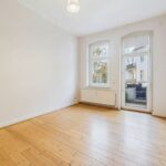 Helles, leerer Raum mit Holzfußboden, großen Fenstern und Zugang zu einem Balkon.