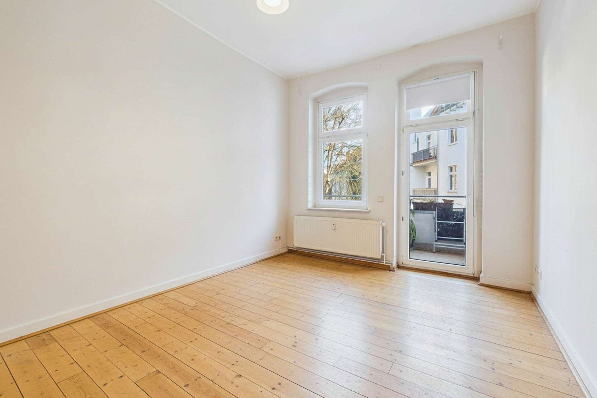 Helles, leerer Raum mit Holzfußboden, großen Fenstern und Zugang zu einem Balkon.
