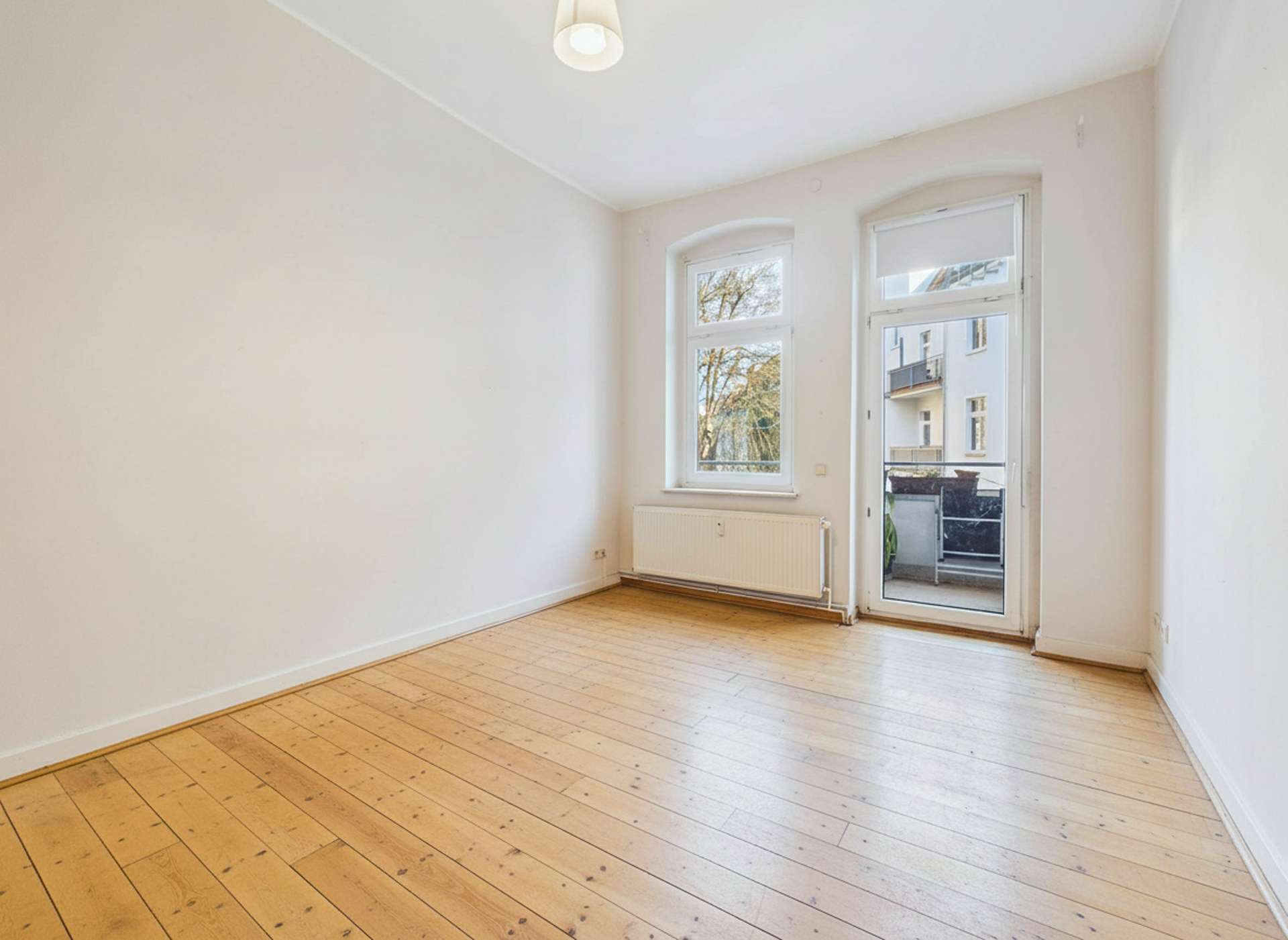 Helles, leerer Raum mit Holzfußboden, großen Fenstern und Zugang zu einem Balkon.