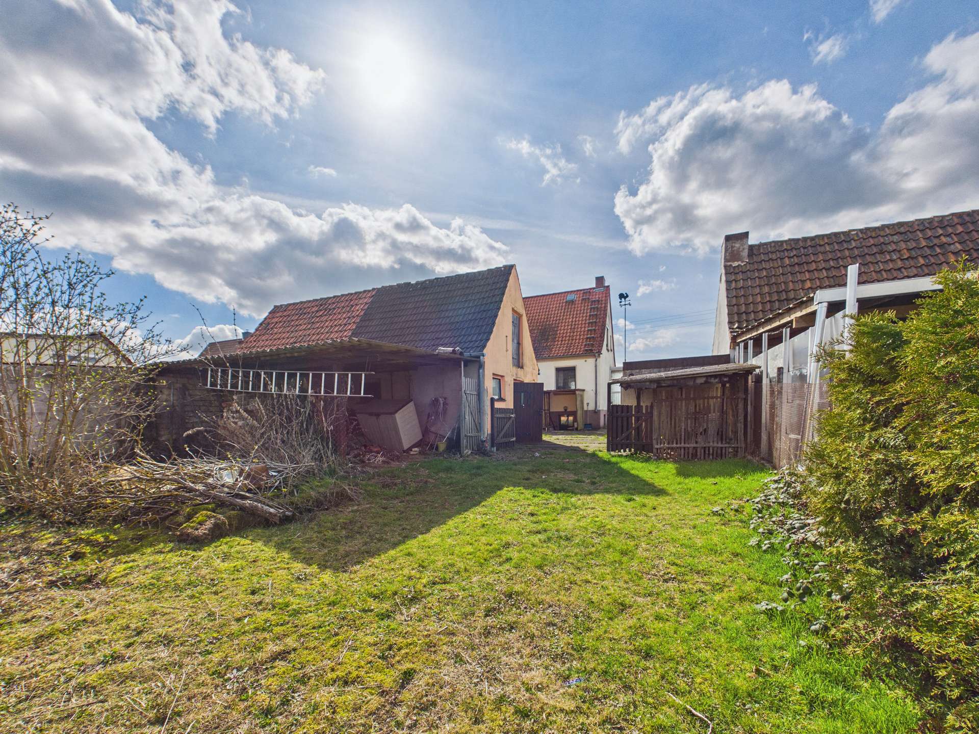 Hinterhof mit zwei Häusern, einer Scheune und grünem Gras, bei sonnigem Himmel mit Wolken.