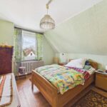 Schlafzimmer mit Dachschräge, Holzbett, buntem Bettzeug und Fenster mit Vorhängen, Blick auf den Himmel.