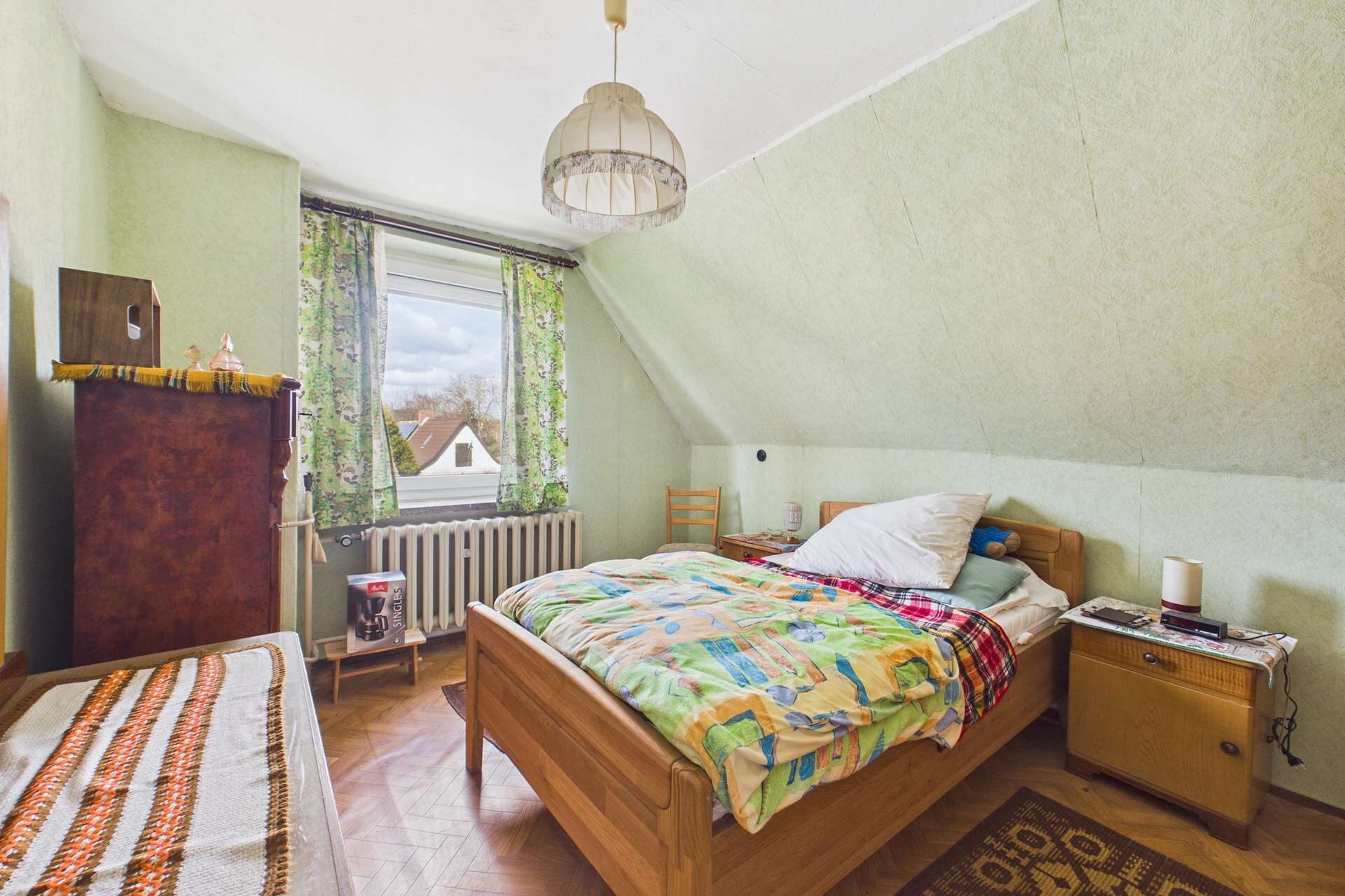 Schlafzimmer mit Dachschräge, Holzbett, buntem Bettzeug und Fenster mit Vorhängen, Blick auf den Himmel.