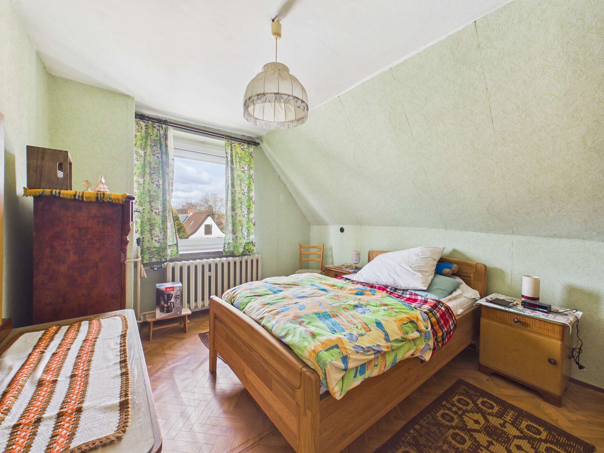 Schlafzimmer mit Dachschräge, Holzbett, buntem Bettzeug und Fenster mit Vorhängen, Blick auf den Himmel.