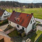 Luftaufnahme eines Hauses mit rotem Dach, umgeben von Grünflächen und einer Straße in ländlicher Umgebung.