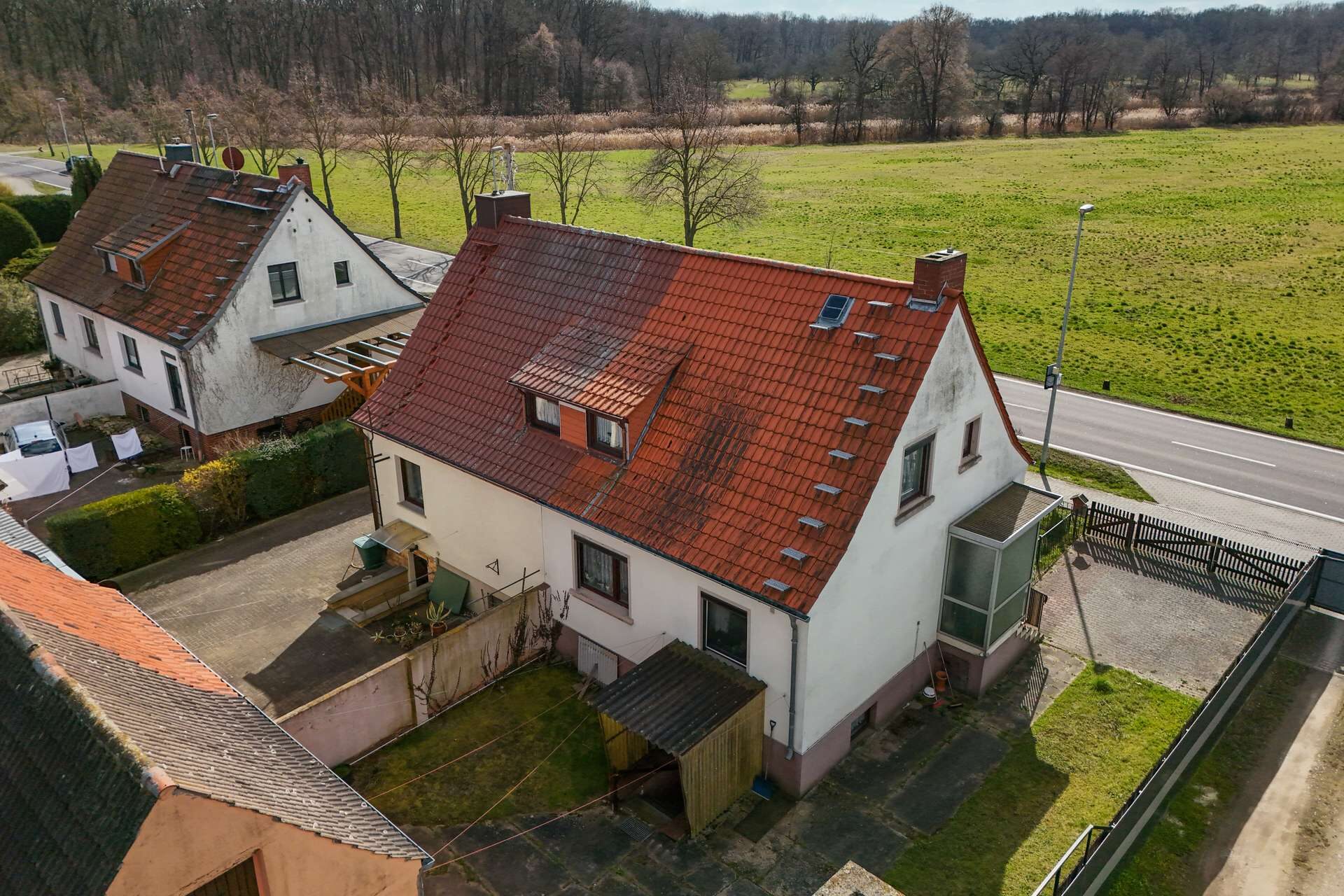 Luftaufnahme eines Hauses mit rotem Dach, umgeben von Grünflächen und einer Straße in ländlicher Umgebung.