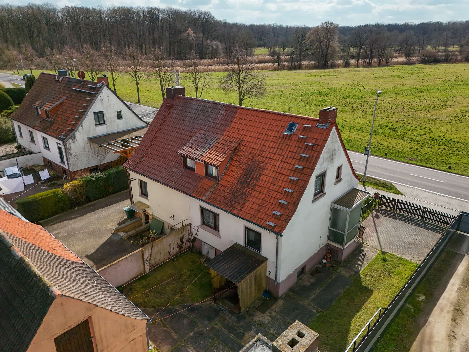 Luftaufnahme eines Hauses mit rotem Dach, umgeben von Grünflächen und einer Straße in ländlicher Umgebung.