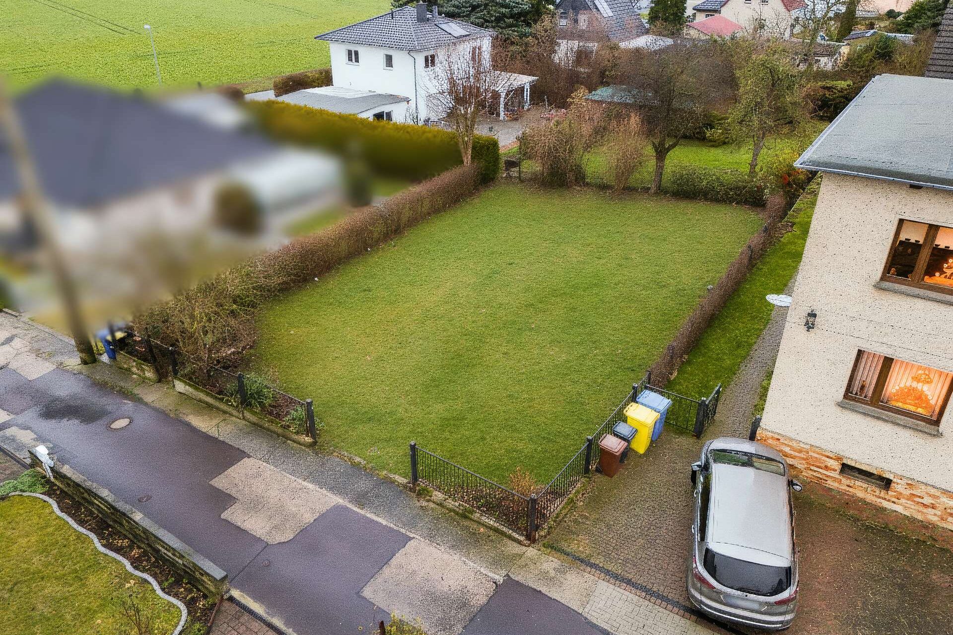 Luftaufnahme eines gepflegten Gartens mit Rasen, umgeben von Hecken und einem Parkplatz neben einem Wohnhaus.