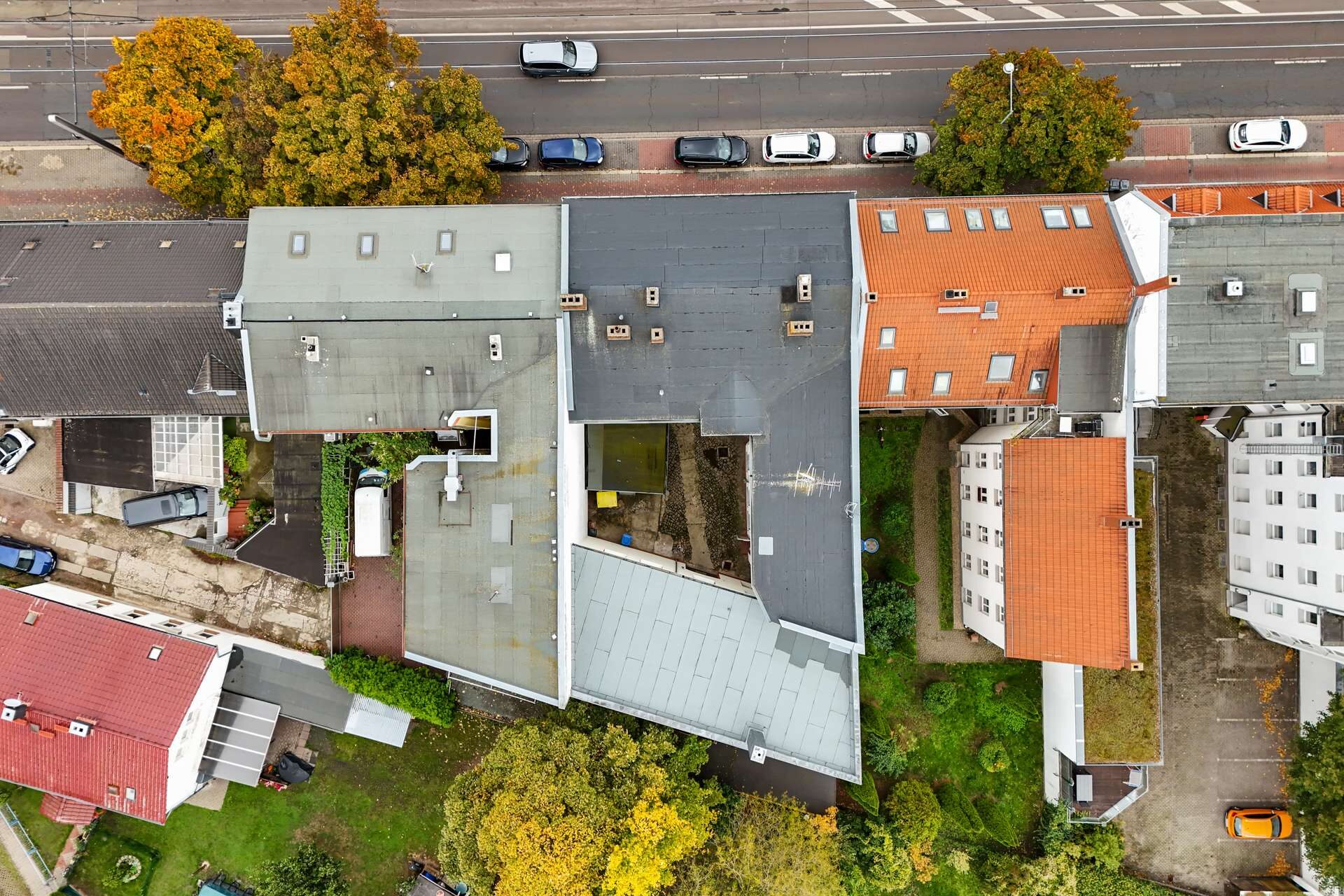 Luftaufnahme eines Wohngebiets mit verschiedenen Dachformen und Bäumen, umgeben von Straßen und parkenden Autos.