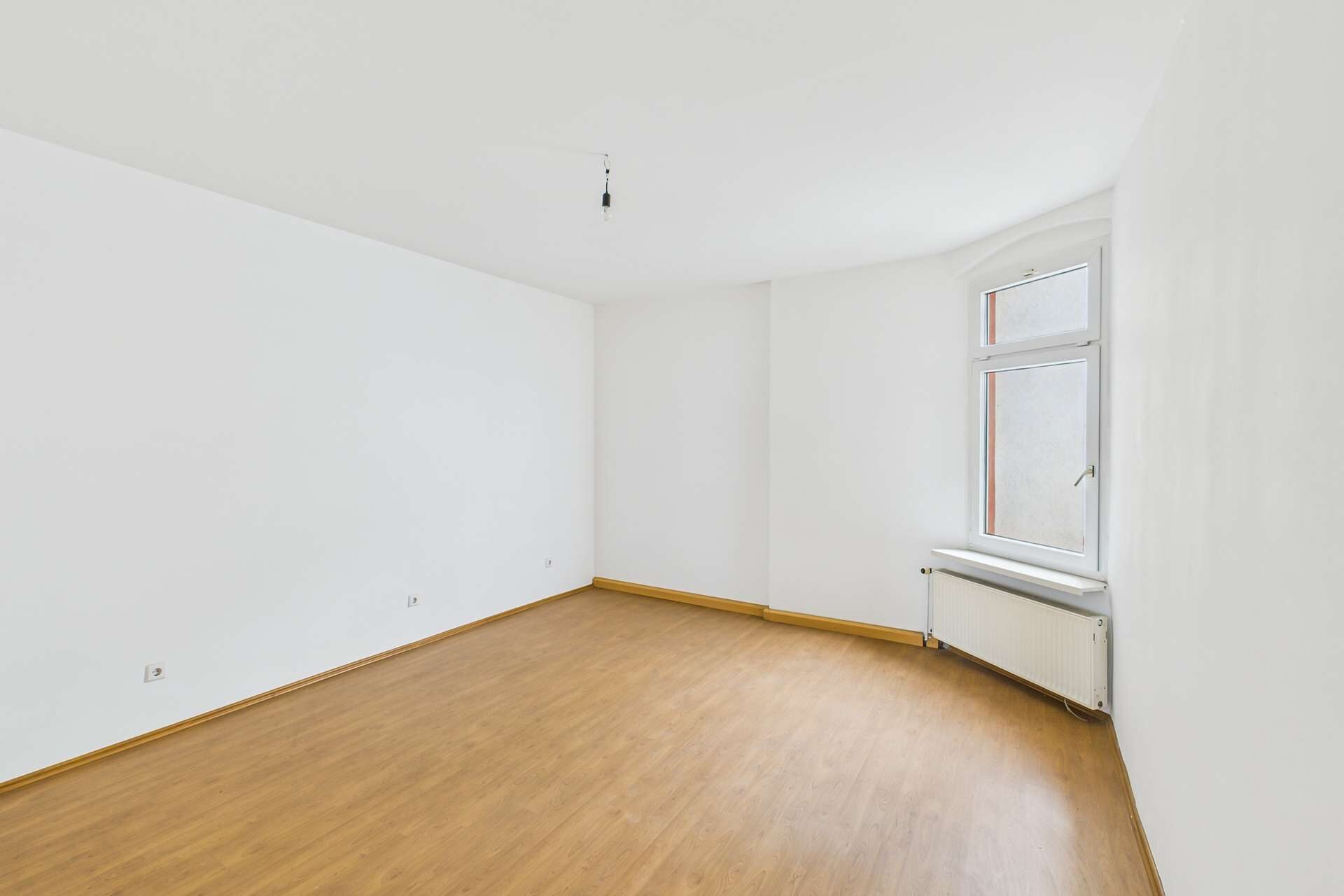 Leerer Raum mit hellem Holzfußboden, weißen Wänden und einem Fenster, das natürliches Licht hereinlässt.