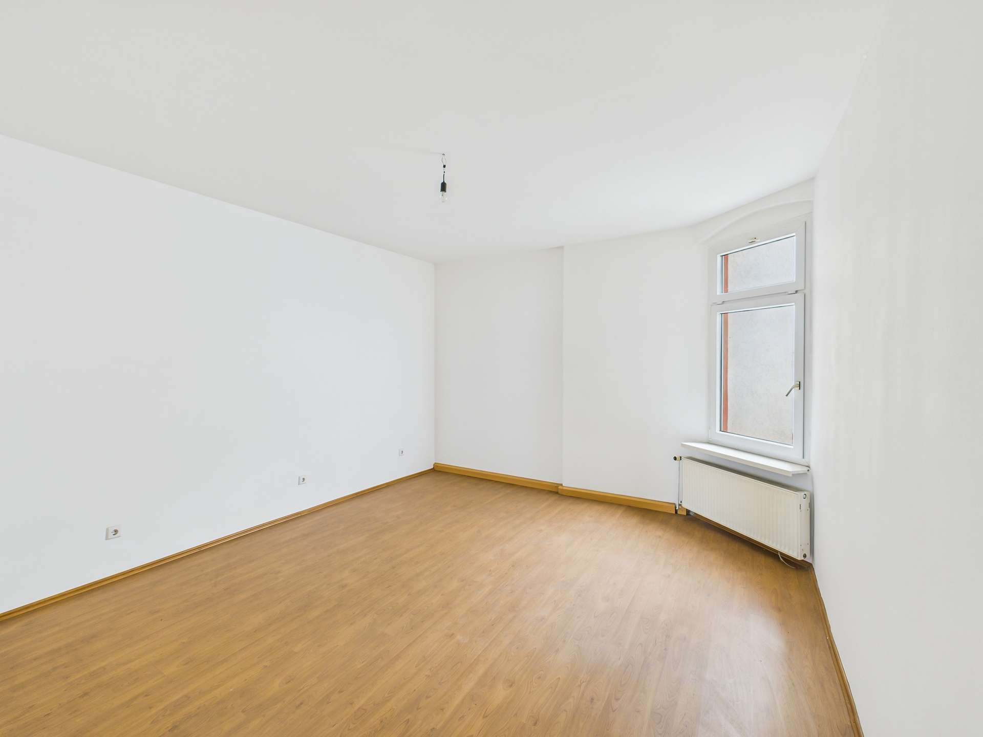 Leerer Raum mit hellem Holzfußboden, weißen Wänden und einem Fenster, das natürliches Licht hereinlässt.