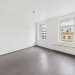 Heller, leerer Raum mit zwei Fenstern und grauem Bodenbelag, weiße Wände und Heizkörper an der Wand.