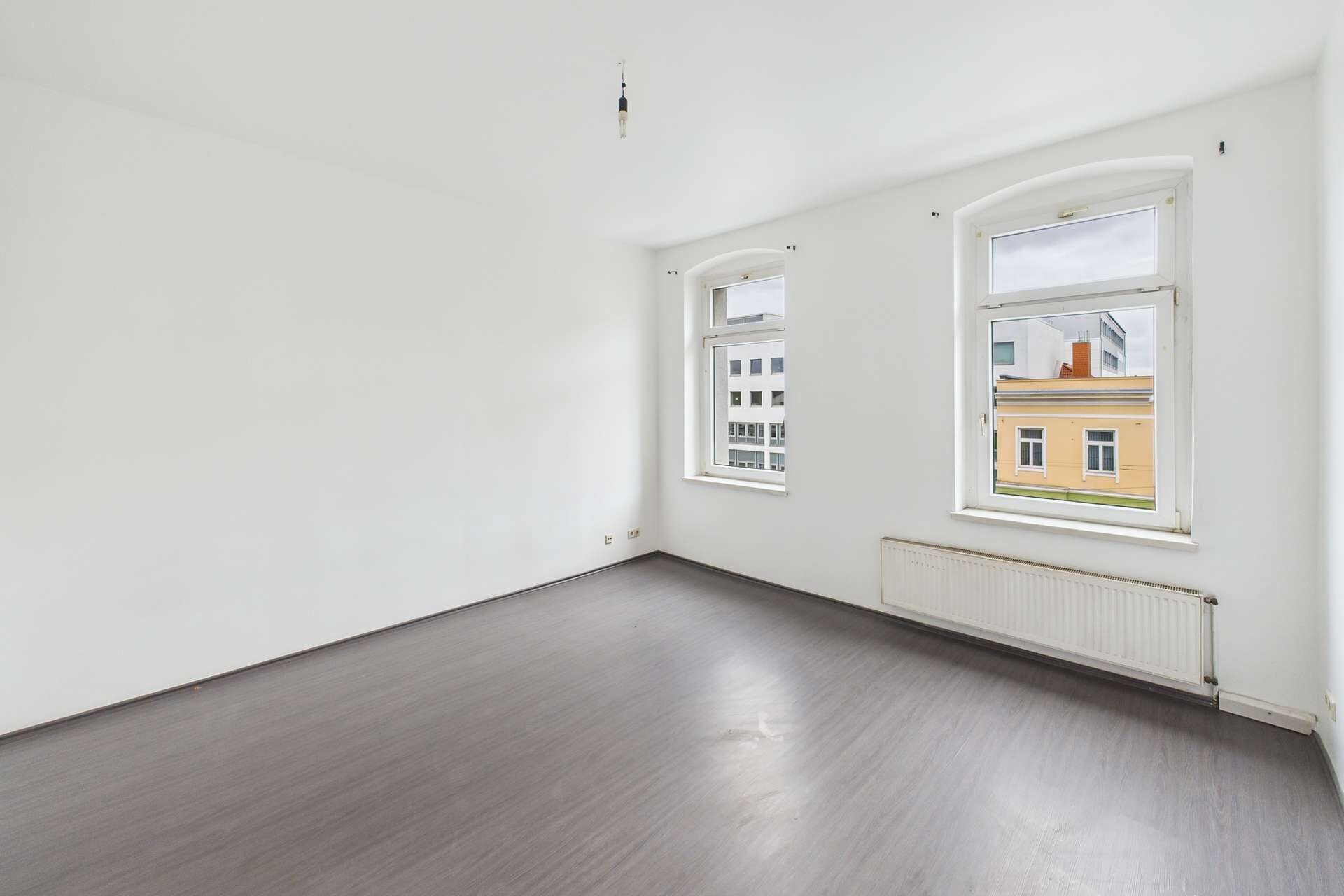 Heller, leerer Raum mit zwei Fenstern und grauem Bodenbelag, weiße Wände und Heizkörper an der Wand.