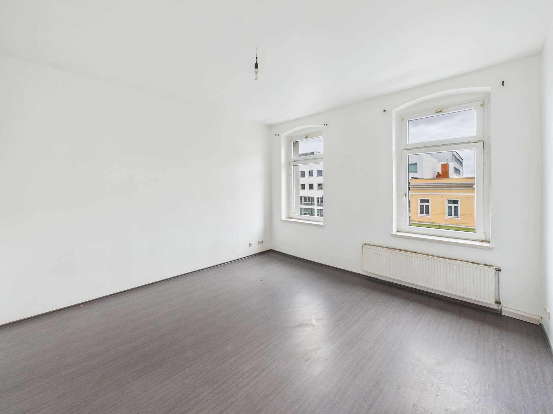 Heller, leerer Raum mit zwei Fenstern und grauem Bodenbelag, weiße Wände und Heizkörper an der Wand.