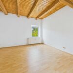 Heller Raum mit Holzdeckenbalken, großem Fenster und hellem Laminatboden.