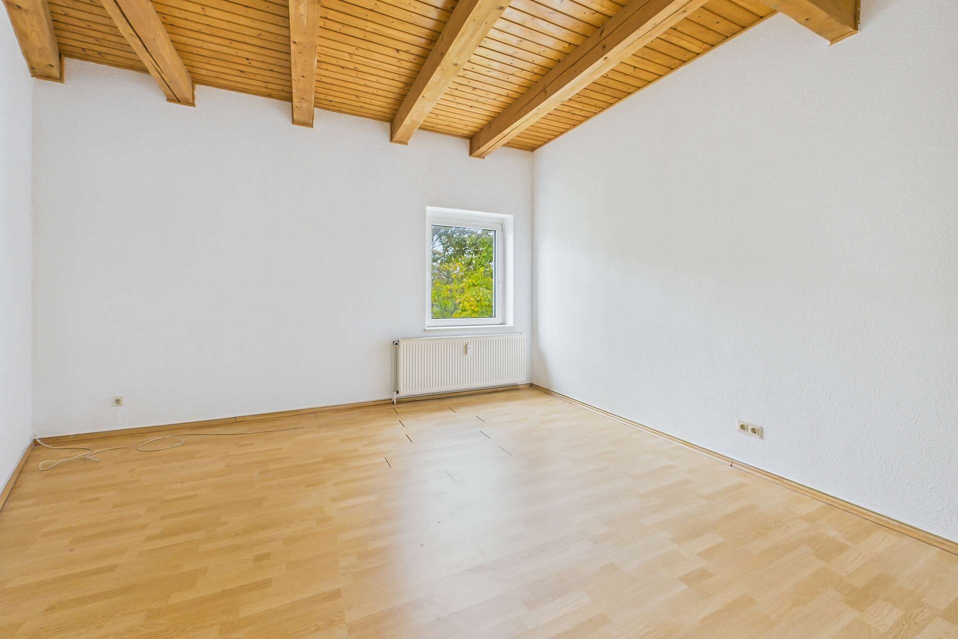 Heller Raum mit Holzdeckenbalken, großem Fenster und hellem Laminatboden.