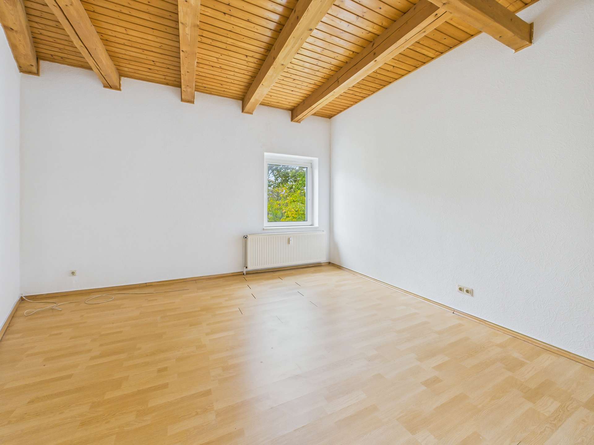 Heller Raum mit Holzdeckenbalken, großem Fenster und hellem Laminatboden.