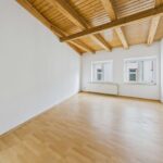 Helle, leere Wohnung mit Holzdecke, zwei Fenstern und einem hellen, lichtdurchfluteten Raum.