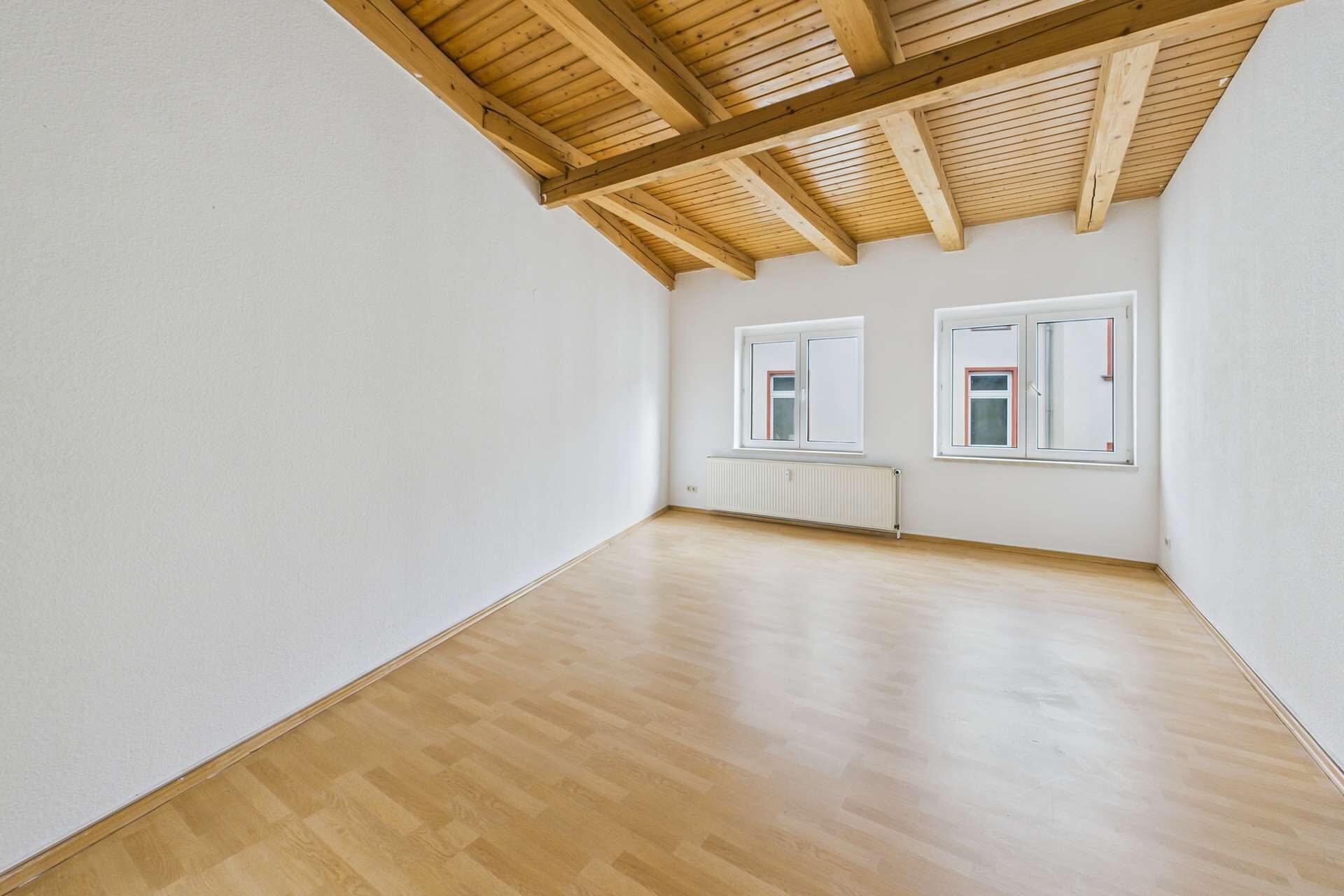 Helle, leere Wohnung mit Holzdecke, zwei Fenstern und einem hellen, lichtdurchfluteten Raum.