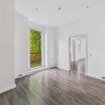 Helles, leerstehendes Zimmer mit Holzfußboden und Fenster, das auf einen Balkon mit Blick ins Grüne führt.