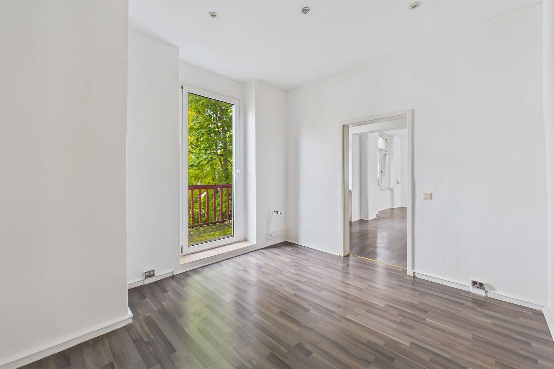 Helles, leerstehendes Zimmer mit Holzfußboden und Fenster, das auf einen Balkon mit Blick ins Grüne führt.