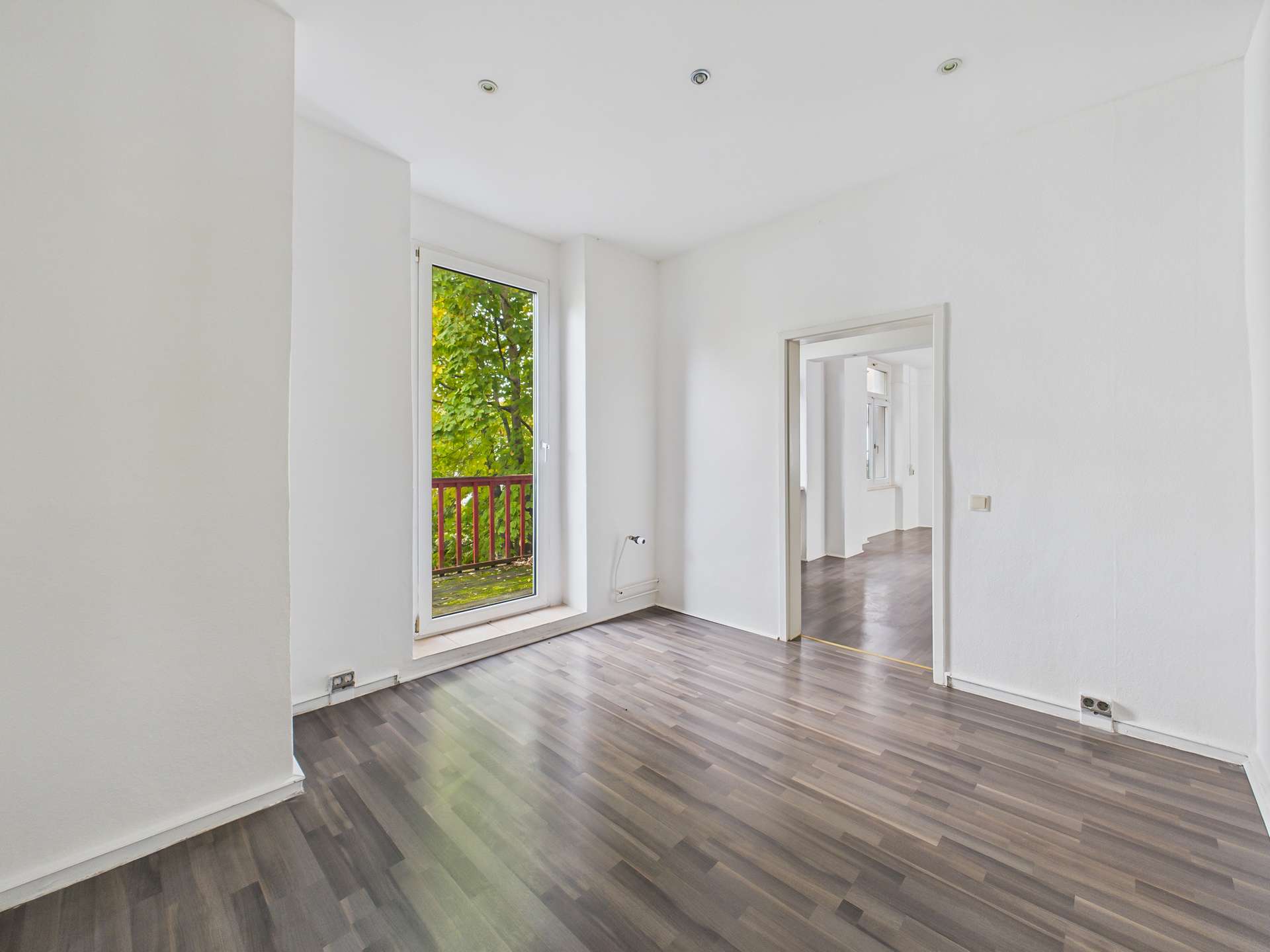 Helles, leerstehendes Zimmer mit Holzfußboden und Fenster, das auf einen Balkon mit Blick ins Grüne führt.
