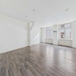 Helle, leere Wohnung mit Holzfußboden und großen Fenstern, die Tageslicht hereinlassen.
