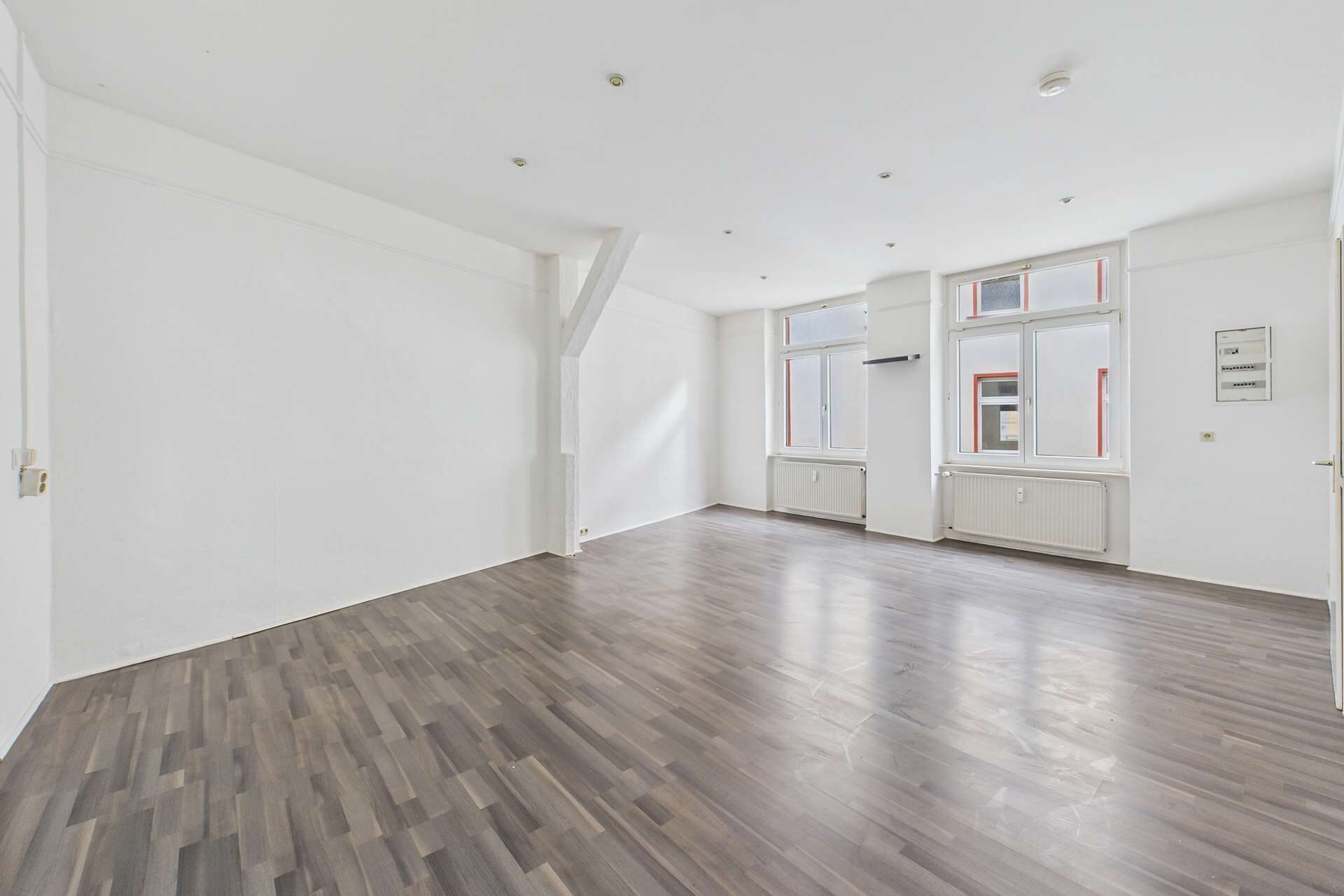 Helle, leere Wohnung mit Holzfußboden und großen Fenstern, die Tageslicht hereinlassen.