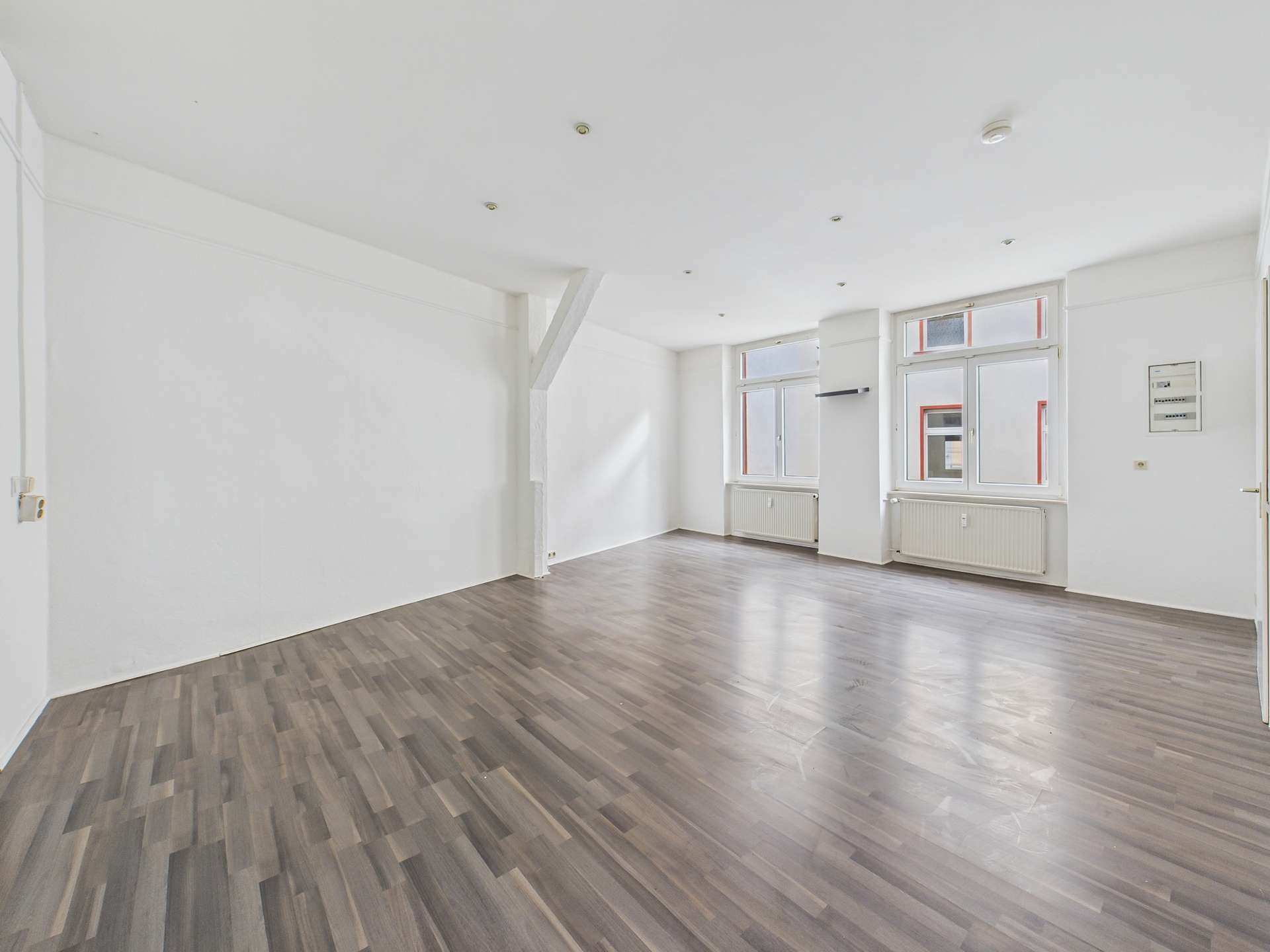 Helle, leere Wohnung mit Holzfußboden und großen Fenstern, die Tageslicht hereinlassen.