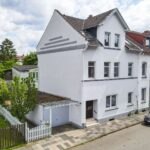 Ein modernes, weißes Wohnhaus mit grauen Akzenten, umgeben von Bäumen und einer gepflasterten Straße.
