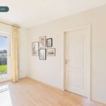 Helle, minimalistische Zimmeransicht mit Fenster, Holzboden und Bilderrahmen an der Wand.