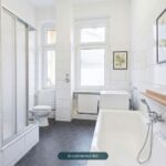 Helles Badezimmer mit Dusche, Badewanne, WC und großen Fenstern, gefliest mit weißen und grauen Kacheln.