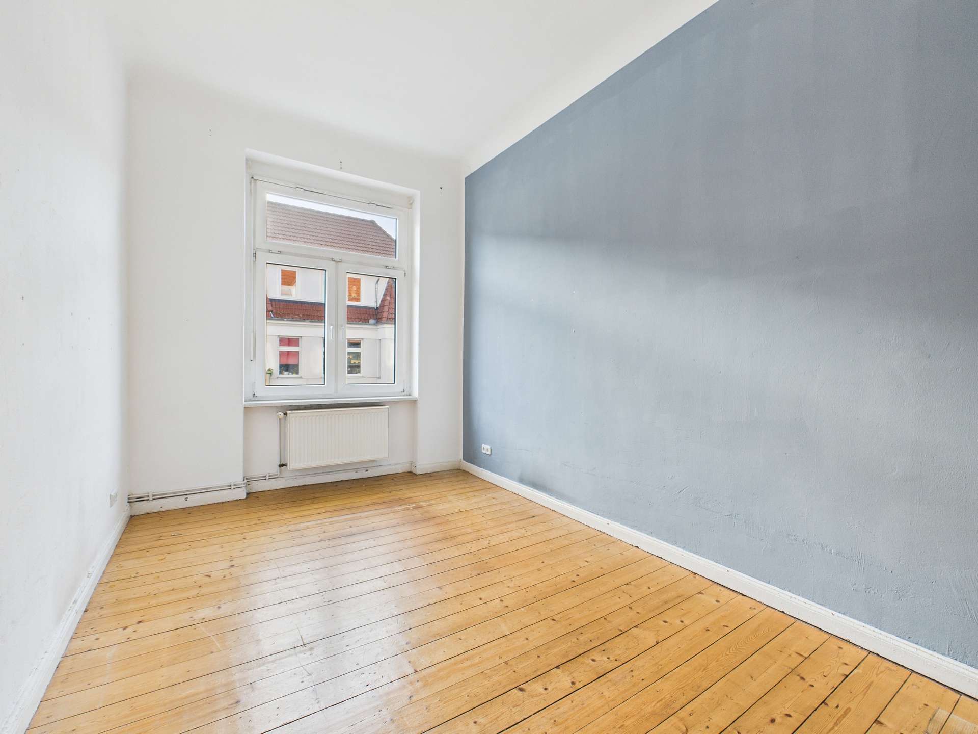 Leerer Raum mit Holzboden, einem Fenster und einer grauen Wand. Natürliches Licht fällt durch das Fenster.