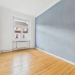 Helle, leere Zimmer mit Holzfußboden und einem grauen Wandabschnitt, Fenster mit Ausblick auf ein Wohngebiet.