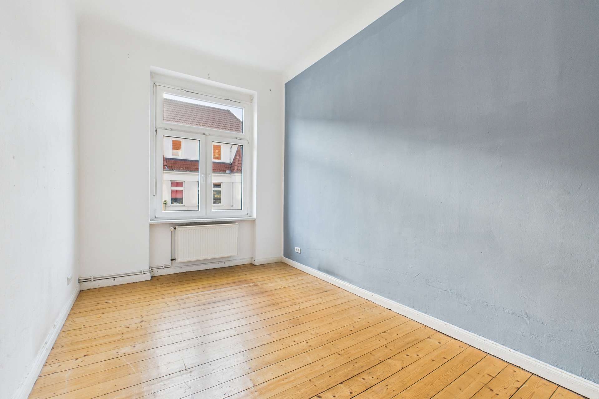 Helle, leere Zimmer mit Holzfußboden und einem grauen Wandabschnitt, Fenster mit Ausblick auf ein Wohngebiet.