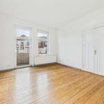 Heller, leerer Raum mit Holzfußboden, zwei Fenstern und Doppeltür, die auf einen Balkon führt.