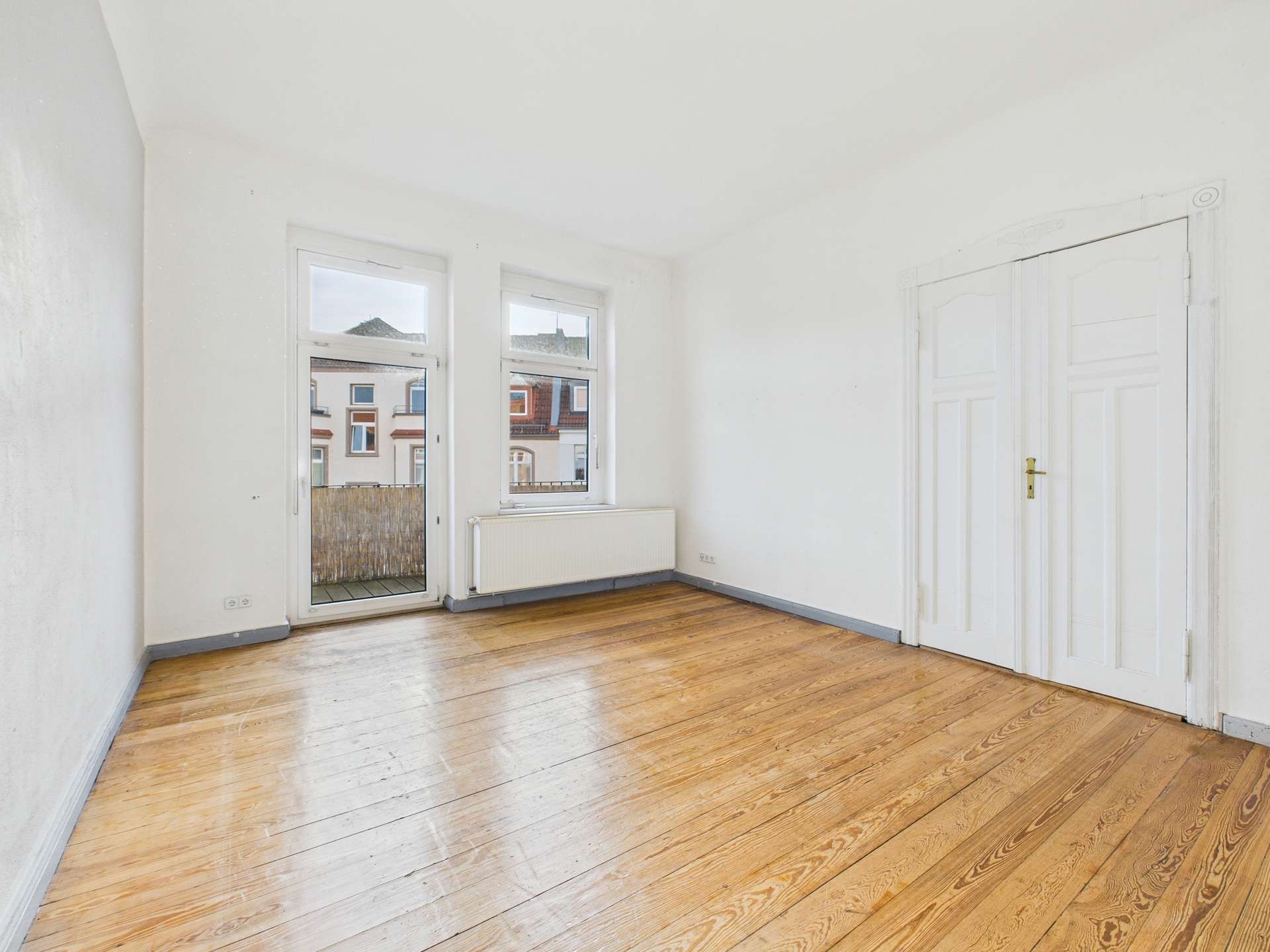 Heller, leerer Raum mit Holzfußboden, zwei Fenstern und Doppeltür, die auf einen Balkon führt.