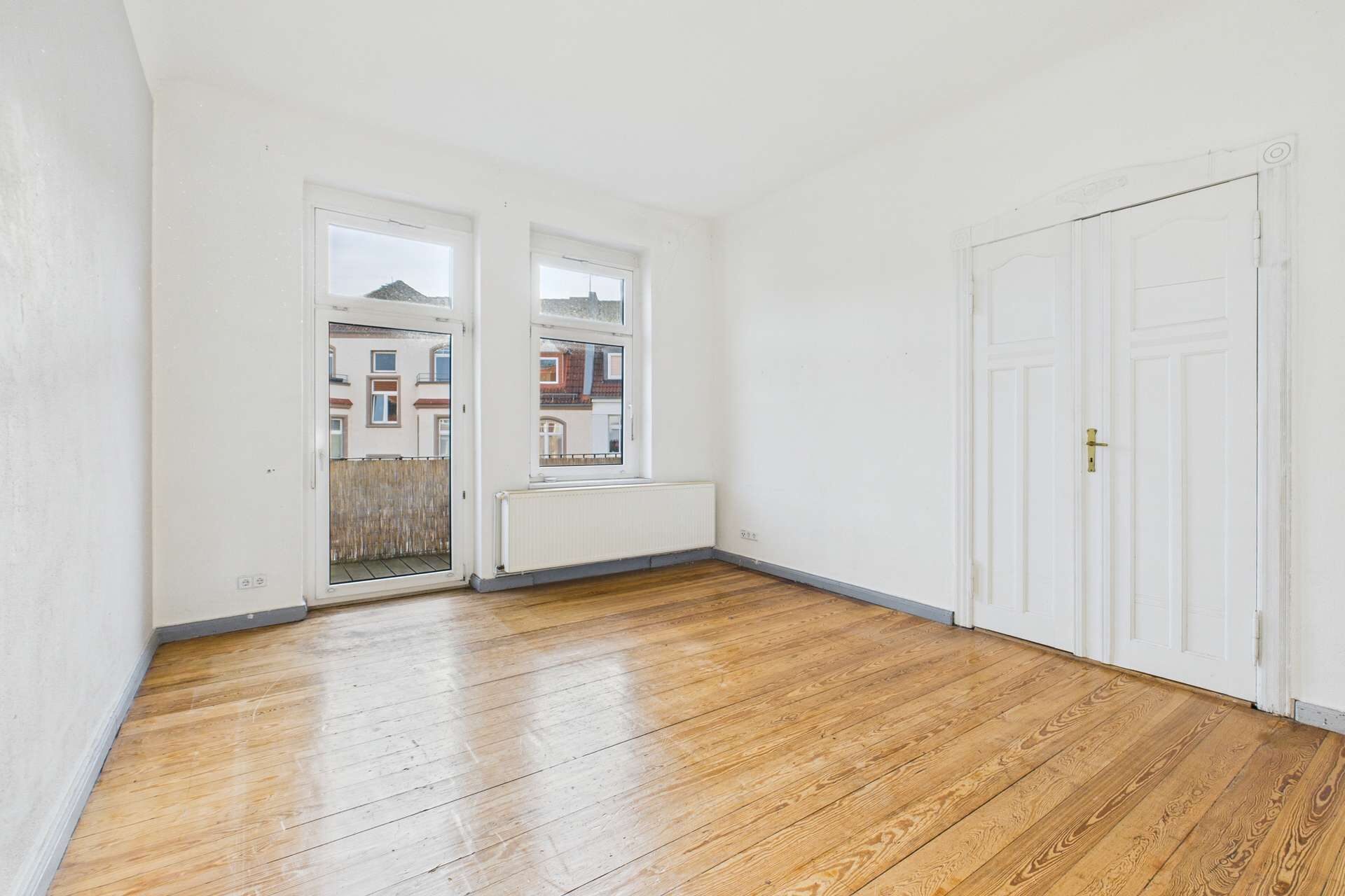Heller, leerer Raum mit Holzfußboden, zwei Fenstern und einer weißen Wand, Blick auf eine Außenansicht.